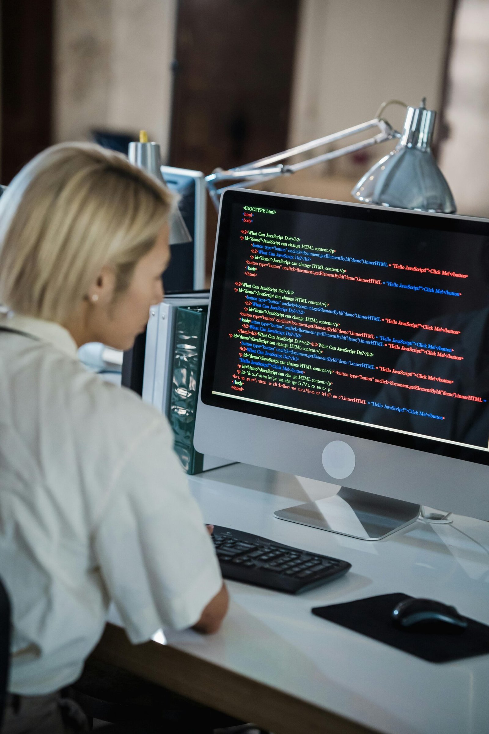 Woman with blonde hair coding on computer in a modern office setting.